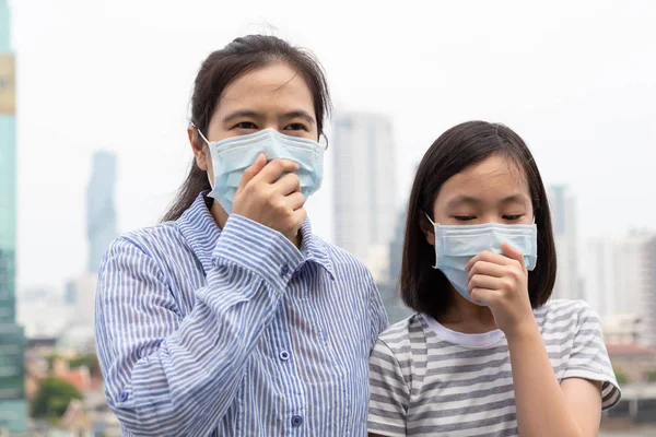 Asian women or mother and daughter suffer from cough with face mask protection,cute child and adult woman wearing face mask because of air pollution in the city building as background,Sick girl with medical mask;concept of pollution,dust allergies