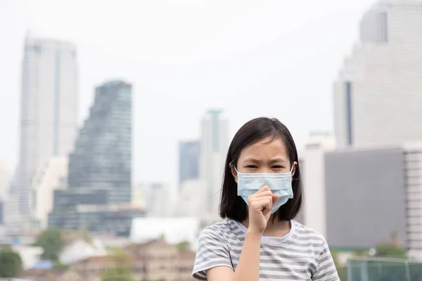 Asiática niña sufre de tos con protección mascarilla, lindo niño con mascarilla debido a la contaminación del aire en el edificio de la ciudad como fondo, niña enferma con máscara médica; concepto de contaminación, alergias al polvo y la salud — Foto de Stock