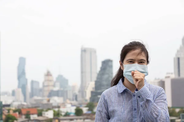 Aziatische jonge vrouw lijdt aan hoest met gezichtsmasker bescherming, zakenvrouw dragen gezichtsmasker vanwege luchtvervuiling in het stadsgebouw als achtergrond, ziek meisje met medisch masker; concept van vervuiling, stof allergieën en gezondheid — Stockfoto