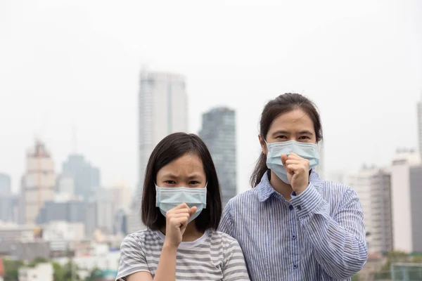 Las mujeres asiáticas o madre e hija sufren de tos con protección contra la máscara facial, lindo niño y mujer adulta que usa mascarilla debido a la contaminación del aire en el edificio de la ciudad como fondo, niña enferma con máscara médica; concepto de contaminación, alergias al polvo — Foto de Stock