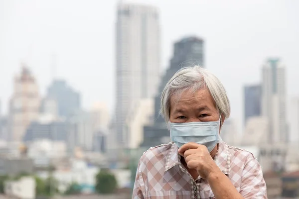 Ασίας ανώτερη γυναίκα υποφέρει από βήχα με προστασία μάσκα προσώπου, ηλικιωμένη γυναίκα που φοράει μάσκα προσώπου λόγω της ατμοσφαιρικής ρύπανσης στο κτίριο της πόλης ως φόντο, άρρωστοι ηλικιωμένοι με ιατρική μάσκα; έννοια της ρύπανσης, των αλλεργιών στη σκόνη και της υγείας — Φωτογραφία Αρχείου
