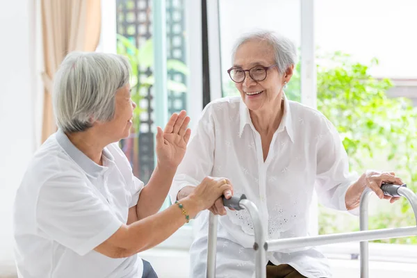 Glückliche zwei asiatische Seniorinnen Händchen haltend für Pflege, Unterstützung und lustige Gespräche, gemeinsame Zeit, alte Menschen lächelnd mit Rollator während der Reha zu Hause, Seniorenfreundschaft, Rentenalter — Stockfoto