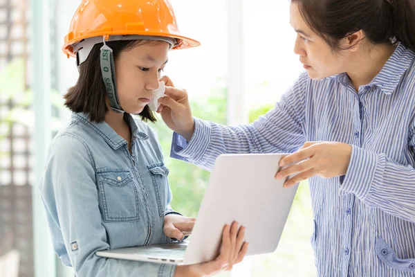 Asian mother using wet wiping face for her daughter,caring woman wipe sweat for girl by tissue paper,cute child with safety helmet and  laptop computer as architect engineer,support care concept
