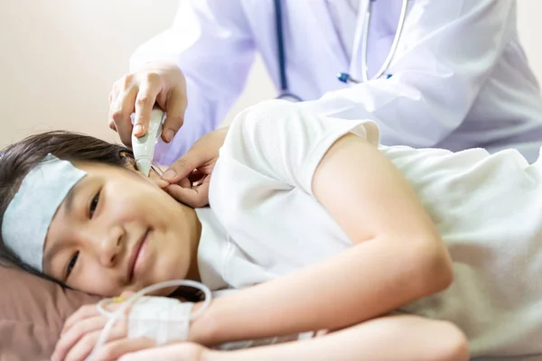 Γιατρός ή νοσοκόμα έλεγχος της θερμοκρασίας του παιδιού ασθενή στο αυτί χρησιμοποιώντας ψηφιακό θερμόμετρο, Ασιατικό μικρό κορίτσι που έχει πυρετό χρησιμοποιώντας κρύο gel τοποθετείται στο μέτωπό της σε ανακούφιση από τον πυρετό, κεφαλαλγία και ασθένεια στο κρεβάτι νοσοκομείο — Φωτογραφία Αρχείου
