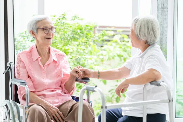 Feliz asiático sênior mulher sentado em cadeira de rodas, irmã ou amigo com walker ter diversão, amigável, mulheres idosas está sorrindo e sua amizade de mãos dadas, falando, rindo em casa, família, conceito de aposentadoria — Fotografia de Stock