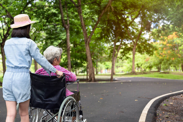 Asian senior woman in wheelchair with little child girl supporting disabled grandparent on walking green nature,grandmother and granddaughter enjoy on street  in summer outdoor park,elderly care,family concept
