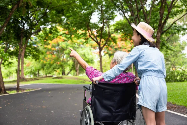 Asiatique femme âgée en fauteuil roulant avec une petite fille enfant soutenant grand-parent handicapé sur la marche nature verte, grand-mère et petite-fille profiter, regarder quelque chose d'intéressant dans le parc extérieur d'été, soins aux personnes âgées, concept de famille — Photo