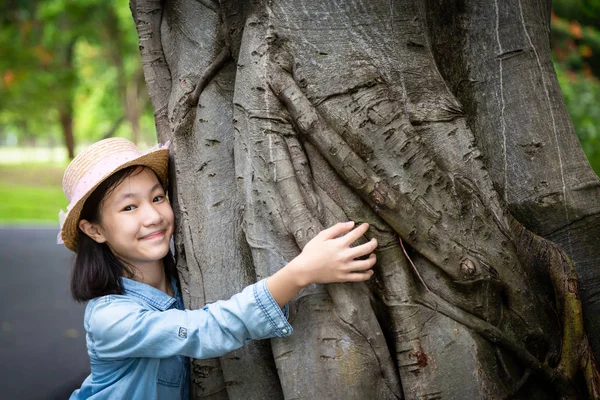 Porträtt av liten flicka i hatt med leende, kramar stora trädstam med armar runt träd och tittar på kameran på utomhusparkering, Asiatiska söta barn med grön natur, lycklig, kärlek, njuta av miljö konceptet — Stockfoto