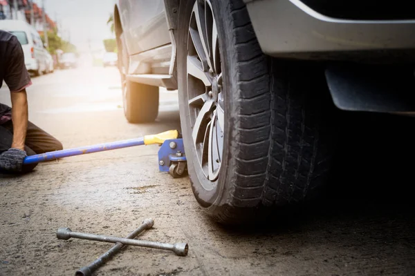 Joven mecánico con guantes, levantando el coche en el gato para la reparación del coche en el aire libre, cambiando neumático pinchado con llave en la carretera, transporte y concepto de viaje —  Fotos de Stock