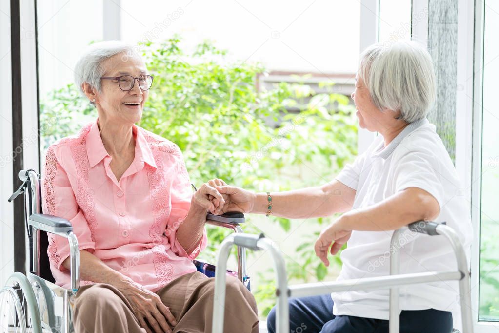 Happy asian senior woman sitting on wheelchair,sister or friend  with walker having fun,friendly,female elderly people is smiling and her friendship holding hands,talking,laughing in house,family,retirement concept