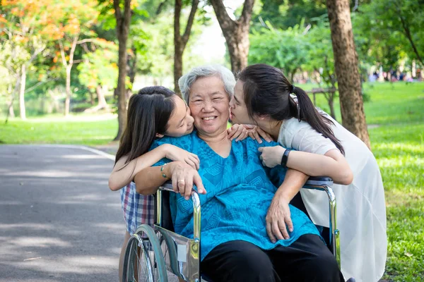 Asiatische Tochter und Enkelin küssen ihre lächelnde ältere Großmutter auf beide Wangen im sommerlichen Outdoor-Park, glückliche Frau, kleine Kinderbetreuung, Liebe, ältere Mutter im Rollstuhl, Familienkonzept — Stockfoto