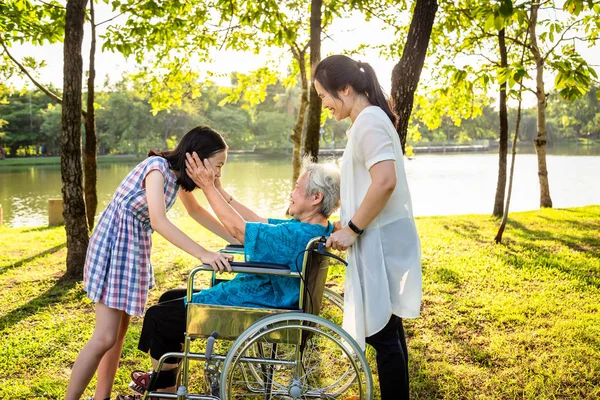 Feliz asiático familia en verano al aire libre parque sonriendo mayor mujer en silla de ruedas mientras su hija riendo y nieta son abrazando su abuela manos tocando mejillas de adorable niño niña, familia, amor, ancianos cuidado concepto — Foto de Stock