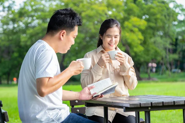 Ευτυχισμένο ζευγάρι της Ασίας απολαμβάνουν, χαμογελούν, μιλούν μαζί, γυναίκα πίνουν καφέ ή τσάι το πρωί, ο σύζυγος κρατήσει ή να διαβάσει ένα βιβλίο Χαλαρώστε στο υπαίθριο πάρκο, νέος άνδρας και γυναίκα το καλοκαίρι πράσινο φύση, οικογένεια, διασκέδαση ιδέα — Φωτογραφία Αρχείου