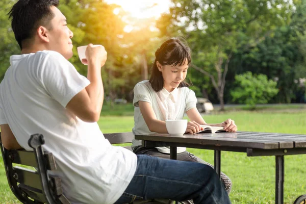 Lycklig asiatisk liten barnflicka eller dotter läser en bok slappna av i utomhusparken, njuta, prata tillsammans, fader Holding Cup varm dricka kaffe eller te i handen på morgonen i sommar grön natur, Familj, fritid, Relationship Concept — Stockfoto