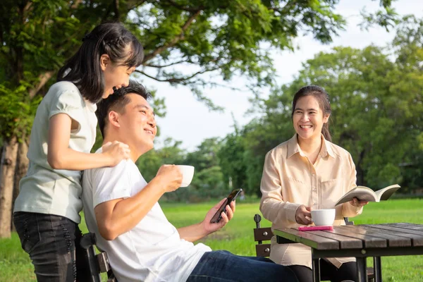 Famiglia asiatica felice, bambina o figlia godono, parlare con il padre, divertirsi insieme, i genitori bevono caffè o tè al mattino, sorridente madre che tiene calda la tazza e leggono un libro rilassarsi nel parco all'aperto, rapporto familiare, concetto di svago — Foto Stock