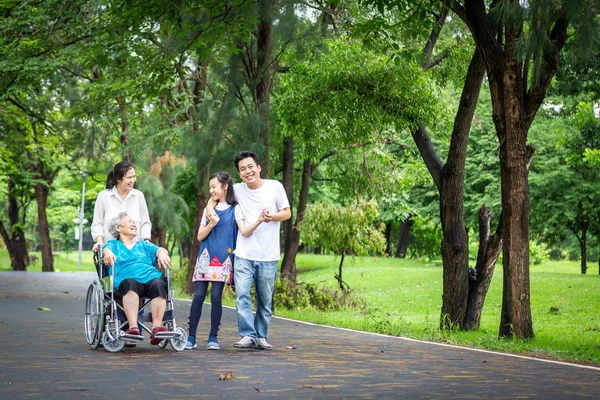 Happy asian family walking, care, talk, child girl or granddaughter and senior grandmother, father, mother having fun, laugh, smile, elderly woman in wheelchair with her family in outdoor park, happy vacation concept — стоковое фото