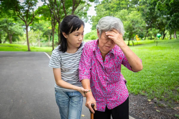Aziatische senior grootmoeder heeft hoofdpijn, aanraken van haar hoofd met haar handen, duizeligheid; zieke ouderen hoge bloeddruk, flauwte, kind meisje of kleindochter zorg, hulp, ondersteuning in de buitenlucht — Stockfoto