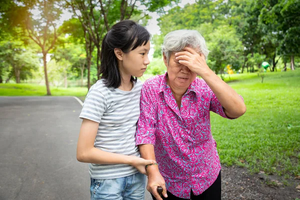 Abuela mayor asiática tiene dolor de cabeza, tocándose la cabeza con las manos, vértigo; mareos; mujer anciana enferma que tiene un golpe de calor, quemaduras solares, se siente débil, niña o nieta cuidado, ayuda, apoyo en el aire libre — Foto de Stock