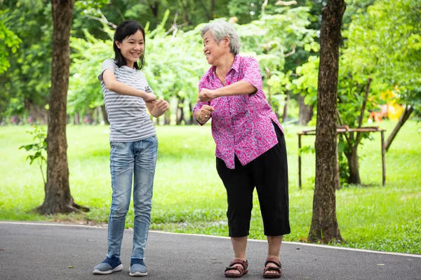 Feliz niña asiática sonriendo y haciendo ejercicio con una anciana en el parque al aire libre, nieta, abuela mayor bailando, relajarse, reír juntos en verano, cuidado de la salud de la familia de la gente, concepto de vacaciones —  Fotos de Stock