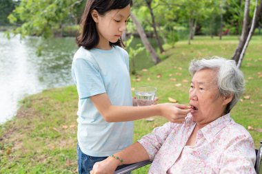 Asyalı çocuk kız veya torunu ilaç hapları veya kapsül el veya parmak ve su, yaşlı büyükanne hap veren, hasta yaşlı kadın alarak, vitamin yeme, antibiyotik, ağrı kesici, tekerlekli sandalye açık parkta besin takviyeleri, sağlık