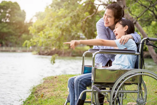 Feliz família asiática, mãe, sorridente filha abraçando, desfrutar, assistindo algo no parque ao ar livre, paciente bonito criança menina rindo falar engraçado com a mãe em cadeira de rodas no verão natureza, reabilitação, relacionamento, conceito de cuidados de saúde — Fotografia de Stock