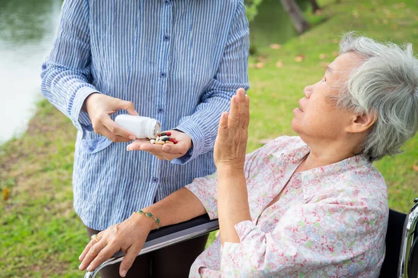 Asiatiska dotter eller vårdgivare hand hälla medicin piller eller kapslar från flaskan, ger piller till senior mor i rullstol utomhus park, sjuk äldre kvinna vägrade att ta medicin, envis, konflikt, hälsovård koncept — Stockfoto