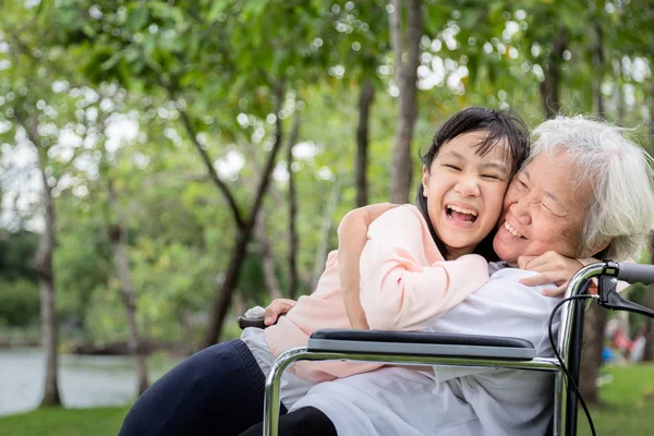 Neta abraçando avó no parque ao ar livre, menina asiática abraço mulher sênior estão passando o tempo juntos, idosos em cadeira de rodas e criança tendo rindo, sorrindo, desfrutando, mãos quentes, cuidado, apoio — Fotografia de Stock