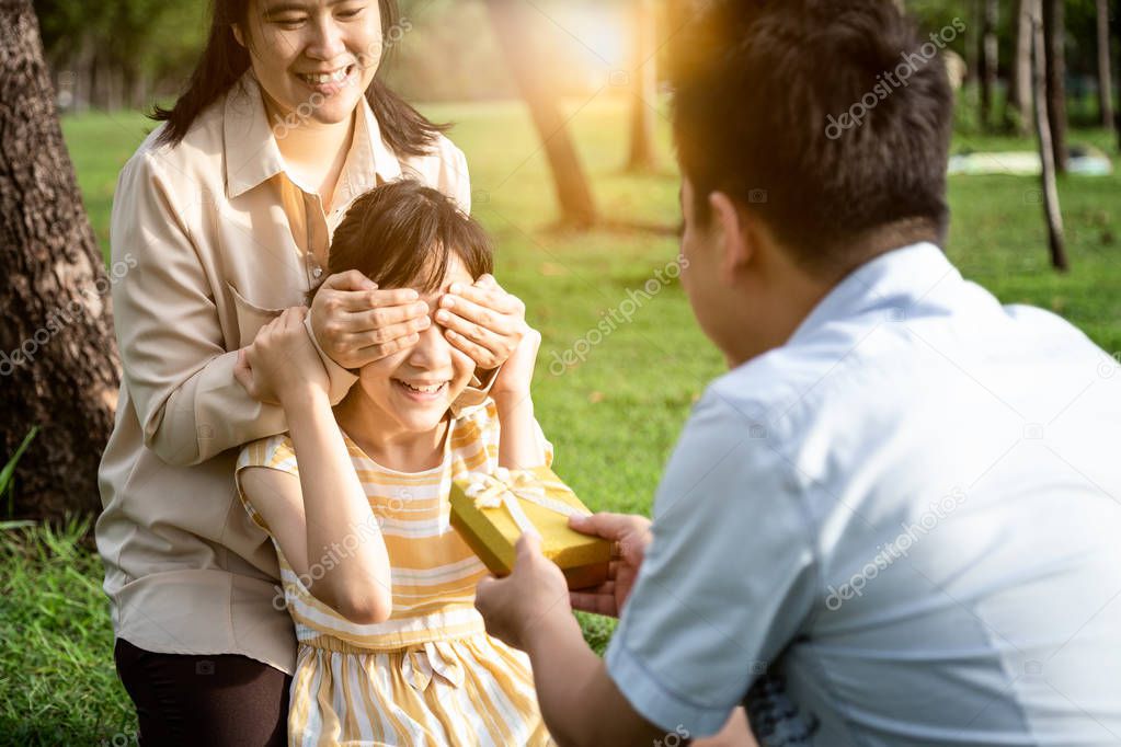 Happy asian family,mother covering eyes of excited daughter and smiling happily in green nature,father giving gift box is congratulating her birthday,parents make surprise for cute little child girl at outdoor park in summer,family relationship conc 