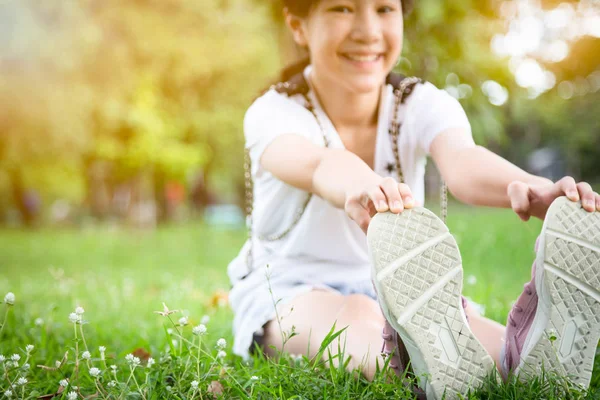 Ázsiai gyönyörű gyermek lány nyújtó lábak ülve parkban, a női tinédzser bemelegítés előtt fut, gyakorlása reggel, mosolygós fitness nő bemelegítés a testét, az egészséges életmód, az egészségügyi koncepció — Stock Fotó