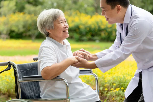 Vänliga manliga asiatiska läkare hälsning återhämta sig, uppmuntrande, tröstande äldre kvinna patient, leende läkare med stetoskop hålla hand äldre människor för att hjälpa vård, stöd på sjukhuset i blomma trädgård, medicinsk, sjukvård, artig koncept — Stockfoto