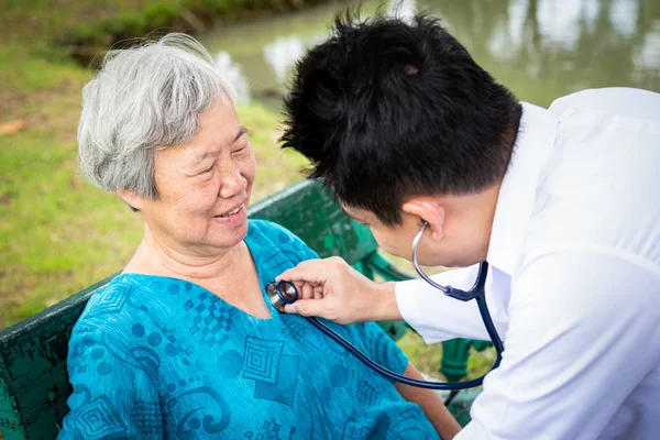 Masculino asiático médico ou filho examinando paciente sênior mulher por estetoscópio, médico de família verificando usando estetoscópio para ouvir respiração de mãe idosa ao ar livre no hospital, exame de saúde geral, conceito de cuidados de saúde — Fotografia de Stock