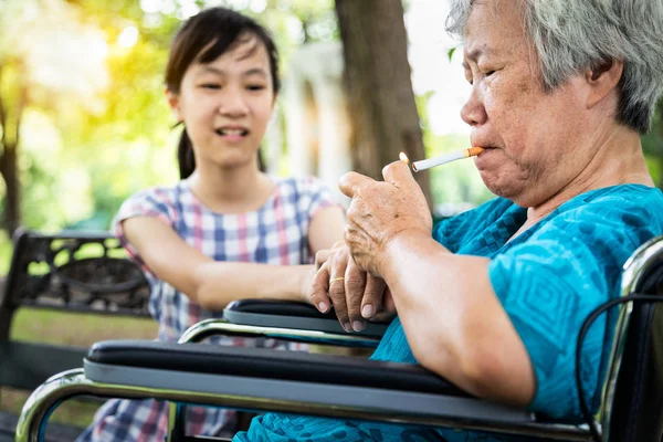 Asiatisches kleines Mädchen, das einen Raucher auffordert, mit dem Rauchen aufzuhören, Rauchen ist gefährlich für die Gesundheit und die Umgebung, Enkelin versucht aufzuhören, warnt oder verbietet ältere Großmutter im Rollstuhl, ältere Frau, die im Park eine Zigarette raucht — Stockfoto