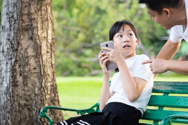 Il padre asiatico chiede alla figlia di smettere di giocare con il suo smartphone mentre è seduto sulla panchina nel parco, l'uomo avverte la bella bambina di non giocare più nel telefono cellulare, smette di usare il cellulare perché ha paura di distruggere la vista — Foto Stock