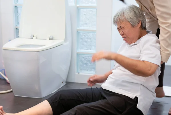 Asian elderly woman on floor in bathroom after falling down because slippery surfaces,caregiver or nurse assisting care,help,support to her,sick senior people fell to the floor in toilet because of dizziness,faint,suffer from illness at nursing home