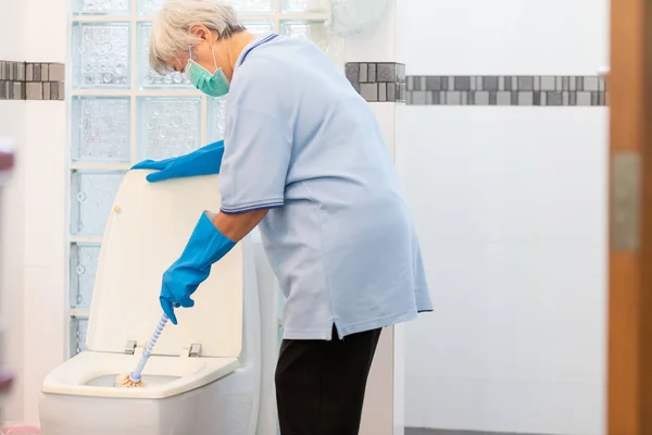 Asian senior woman in blue glove with medical mask cleaning toilet bowl using brush and detergent,female elderly housekeeper with face mask protection clening,wash the bathroom,household duties,house cleaning concept — Stock Photo, Image
