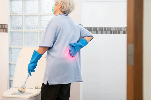 Asian senior woman in blue glove,cleaning toilet bowl,female elderly housekeeper hands touching back pain having backache,muscle,hip pain or waist pain while housework,wash the bathroom at home,health problems concept — Stock Photo, Image