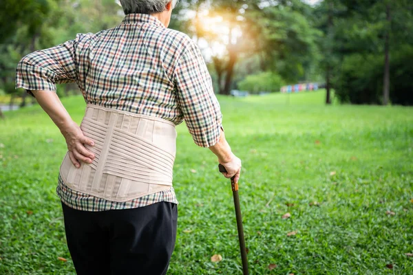 Азиатская старшая женщина с ремнем поддержки спины для защиты ее спины боль, мышечная травма, пожилые люди рука касаясь на бедре с болями в пояснице, боли в спине, страдает от боли в пояснице на открытом воздухе природы в парке — стоковое фото