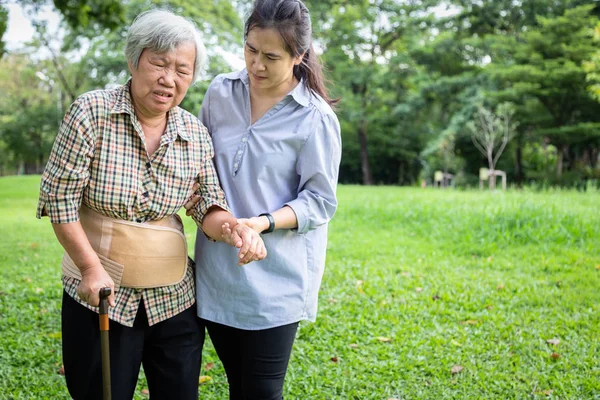 Asiático sênior mãe dor nas costas usar cinto de apoio para proteger a dor nas costas, lesão muscular, meia-idade filha ou assistente de assistência ajuda, apoiar a mulher idosa sentir dor nas costas súbita enquanto caminha no parque — Fotografia de Stock