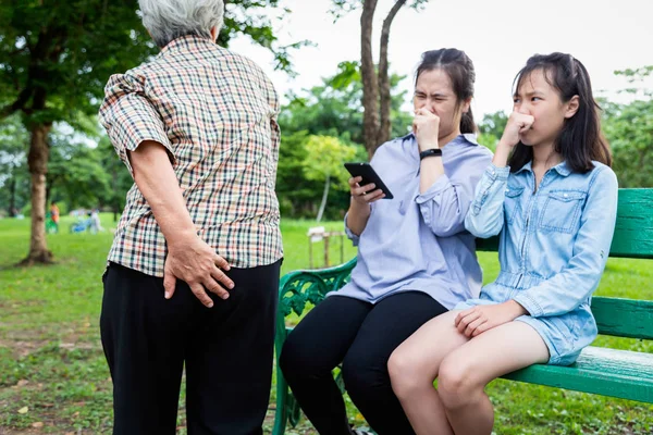Asiatisk mor och dotter klämmer näsan med fingrar, något stinkande och äckligt, senior mormor håller sin rumpa, äldre människor fiser gör en dålig lukt lider av gasbildning och gasbildning i mag-tarmkanalen i parken — Stockfoto