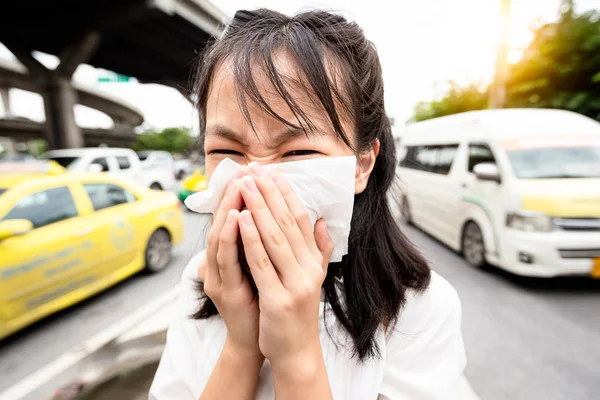 Retrato de primer plano de niña sonándose la nariz en pañuelo de papel, mujer asiática estornudando en un pañuelo en la calle de la ciudad, humos tóxicos del coche, mal olor, concepto de contaminación del aire, alergias al polvo, cuidado de la salud — Foto de Stock