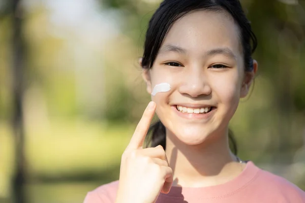 Happy smiling asian child girl apply sunscreen lotion on the cheek,protect her face,schoolgirl using skin care product for sensitive skin,sun block day cream on her delicate facial skin in sunny da