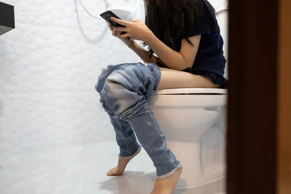 有便秘的亚洲女孩 在浴室里玩网络游戏的女人或社交媒体成瘾的女人 长时间坐在马桶上的人都会导致痔疮 生活方式 — 图库照片