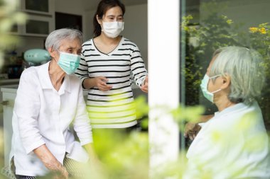 Yüzünde tıbbi maskeler olan Asyalı kadın ve yaşlı bir kadın, mutlu bir şekilde konuşan kız, evde yaşlı annesini ziyaret ediyor, birbirlerine yakın oldukları sırada koruyucu bir maske takıyor, yeni normal bir hayat sürüyor.