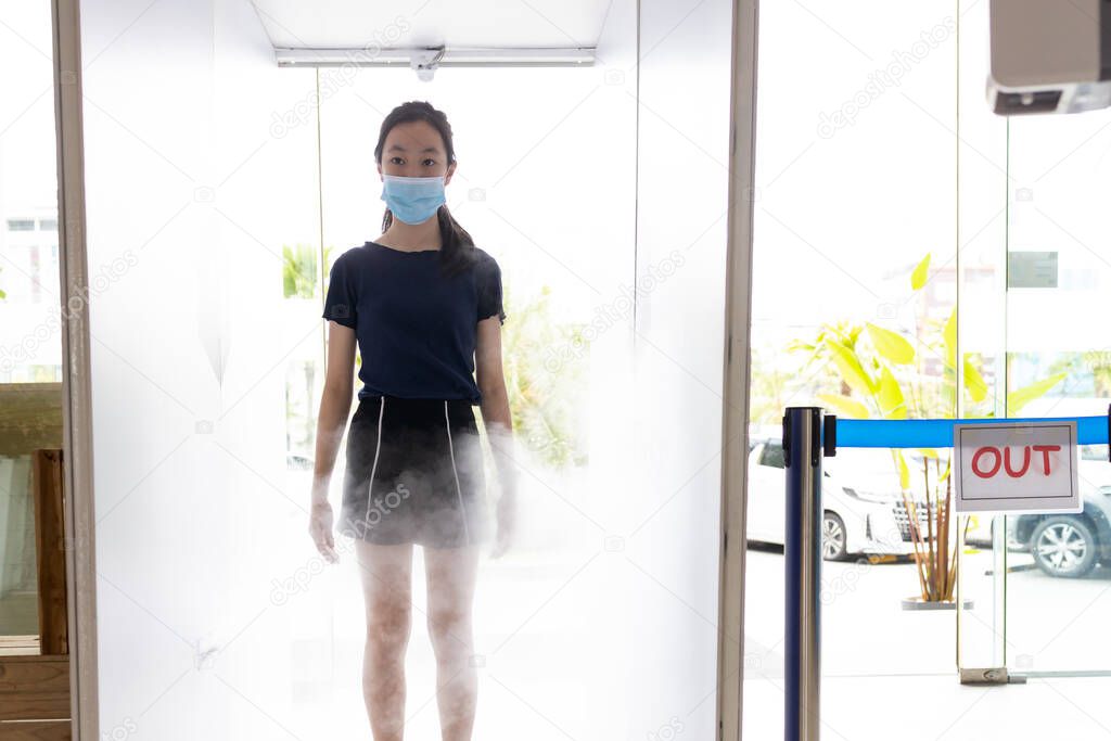 Asian child girl wear face mask,automatic disinfection tunnel is spraying on body for disinfecting at gate check point for COVID-19,measures to prevent,process of cleaning,new normal under Coronavirus