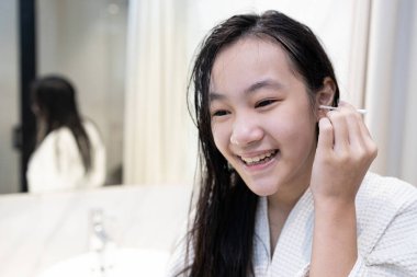 Asian teenager girl cleaning the external ear canal with a cotton swab,smiling young woman in white bathrobe,using cotton bud to wipe ears to dry after shower in bathroom,removing ear wax,health care clipart