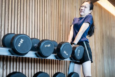 Yaramaz genç kız spor salonundaki halterlerin üzerinde büyük halterleri kaldırmaya çalışıyor, ağırlık kaldırma egzersizlerini, zarar görme veya yaralanma olasılığını, kaza geçirme riskini ve akut kas ağrısını gösteriyor.
