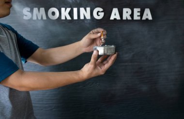Hands of asian man holding a ashtray and cigarette butts,smoker blowing smoke out his mouth while smoking cigarette in smoking area zone,designated smoking area text on the black wood background clipart