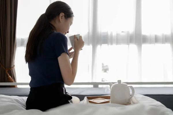 亚洲女孩早上坐在床上喝着热饮料 女人端着一杯草茶 闭着眼睛吸着臭气笑着 香喷喷的热茶 在家里尽情地喝着 — 图库照片