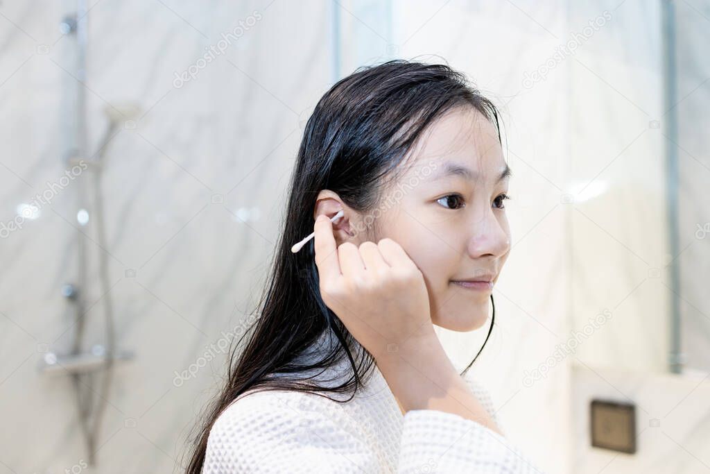 Asian child girl cleaning the external ear canal with a cotton swab,beautiful girl in white bathrobe,using cotton bud to wipe ears to dry after bathing and washing hair or removing earwax,care,hygiene