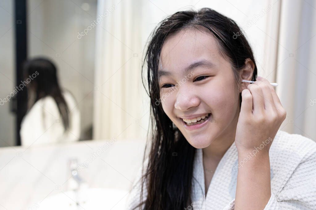 Asian teenager girl cleaning the external ear canal with a cotton swab,smiling young woman in white bathrobe,using cotton bud to wipe ears to dry after shower in bathroom,removing ear wax,health care